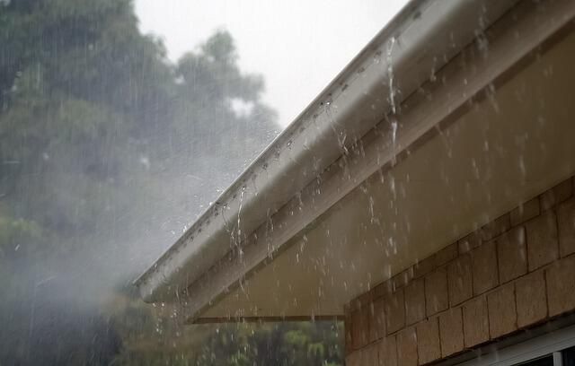 Rain falling from a light-colored gutter on a brown shingled roof, overcast sky.