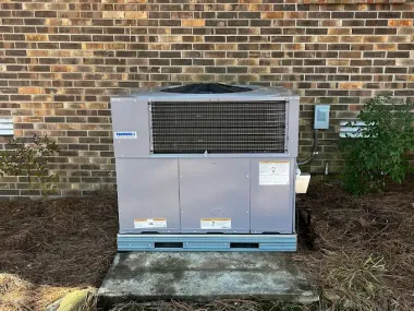 A large, grey HVAC unit sits on a concrete pad in front of a brick wall.