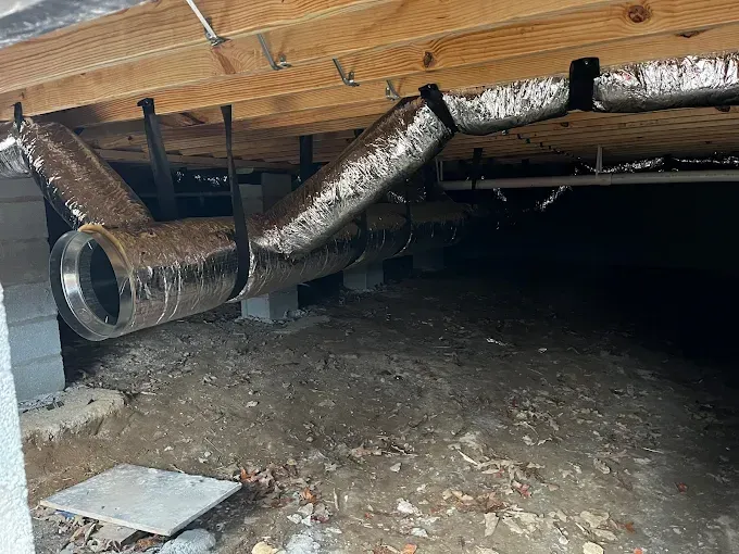 Underneath a house, metallic HVAC ductwork hangs between wooden beams, supported by straps.