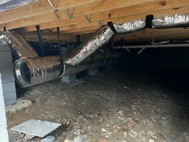 Underneath a house, metallic HVAC ductwork hangs between wooden beams, supported by straps.