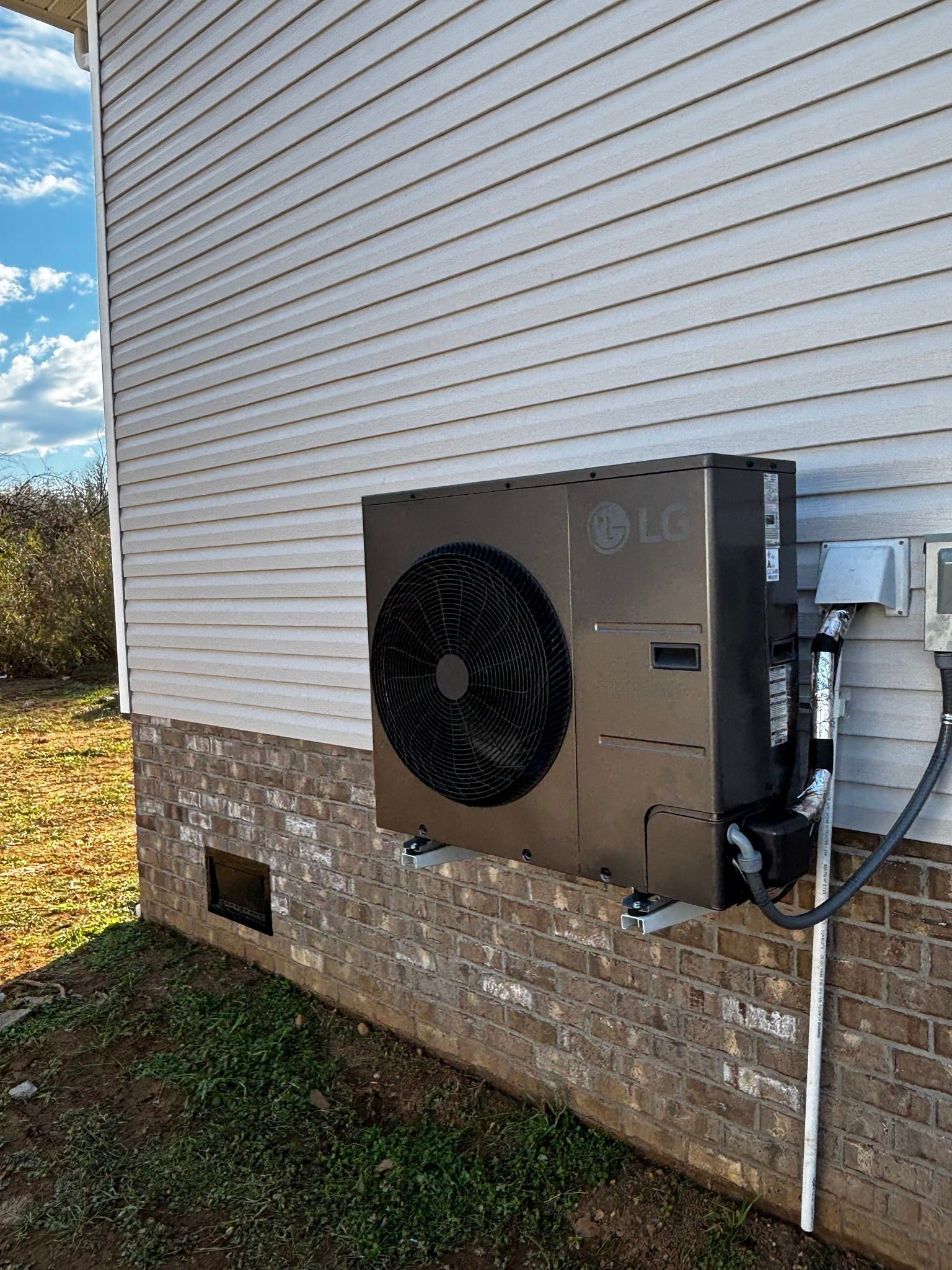 Exterior view of an LG heat pump mounted on a brick and siding wall; overcast sky.
