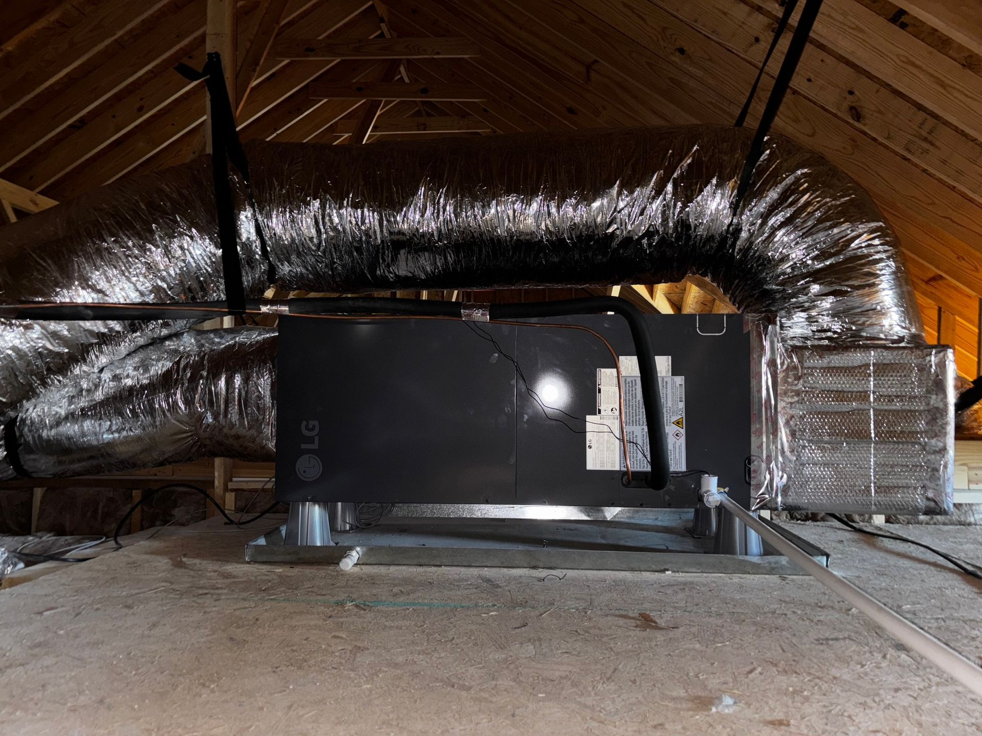 HVAC unit with ductwork in an attic. Silver flexible ducting, black unit, wood beams, and insulation.