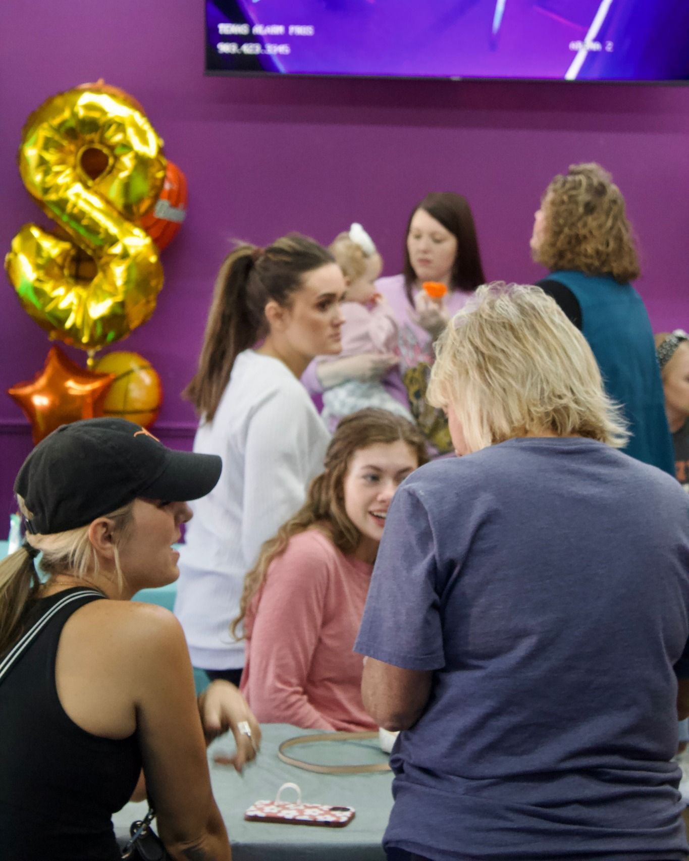 A group of people are gathered around a table with balloons in the background that have the number 8 on them