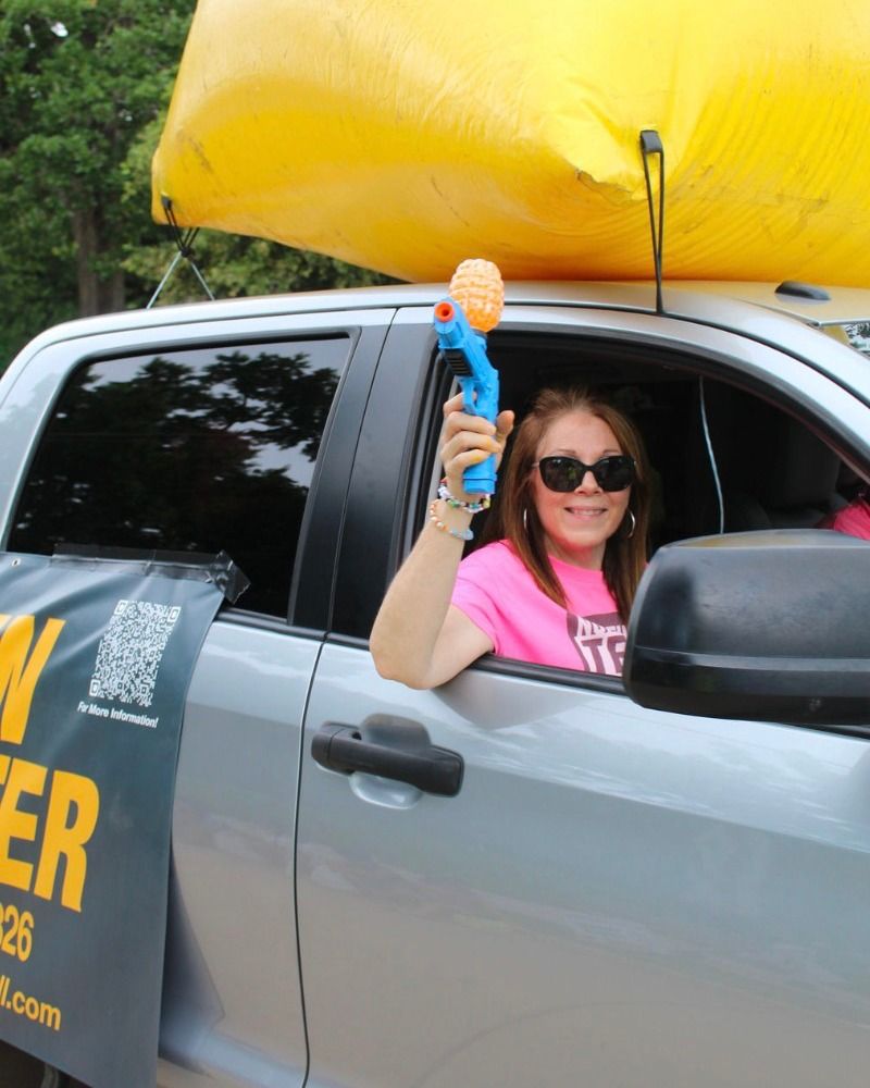 Smiling woman in sunglasses holding a toy blaster from a car window.