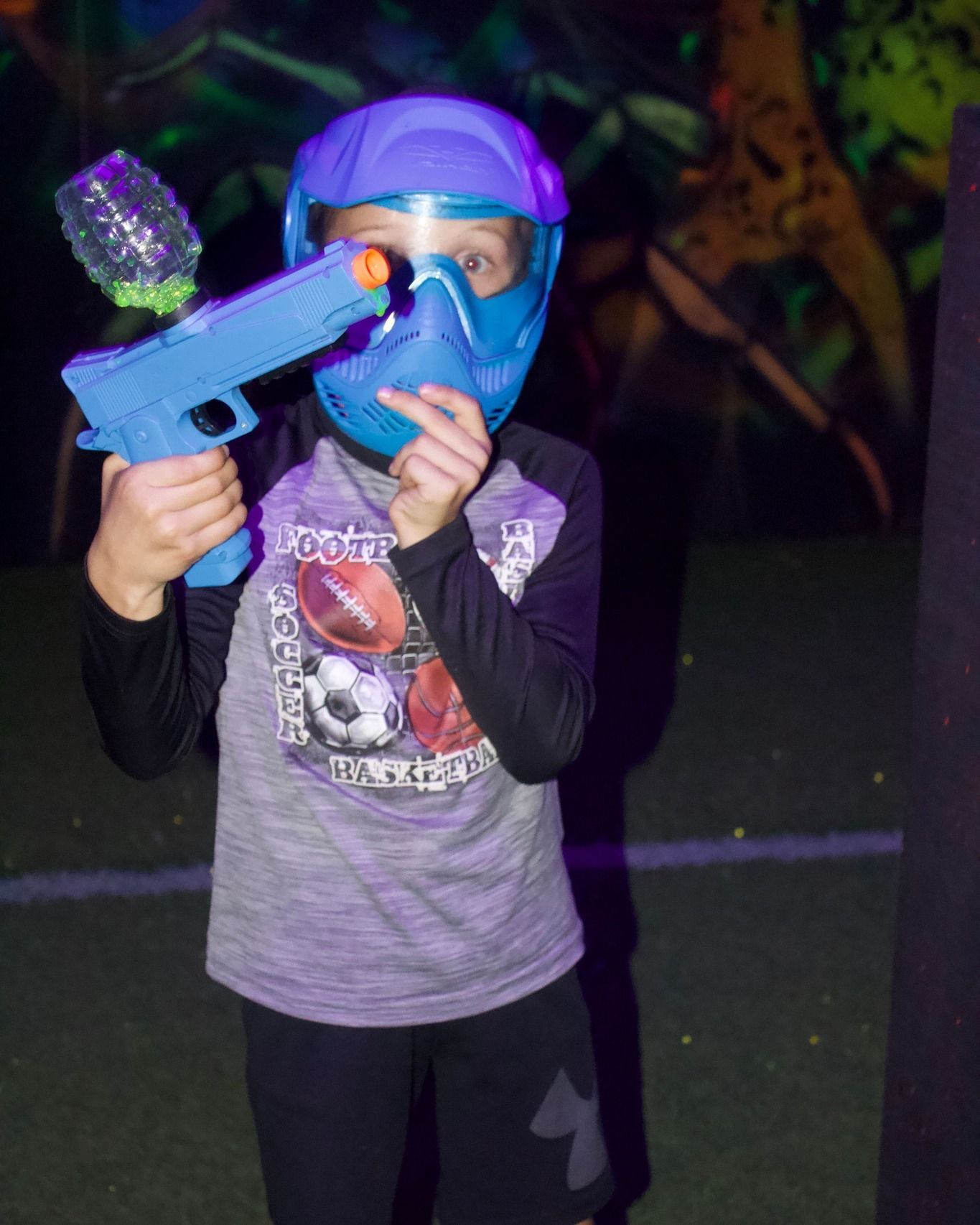Child in a protective mask aiming a toy blaster in a blacklight arena.