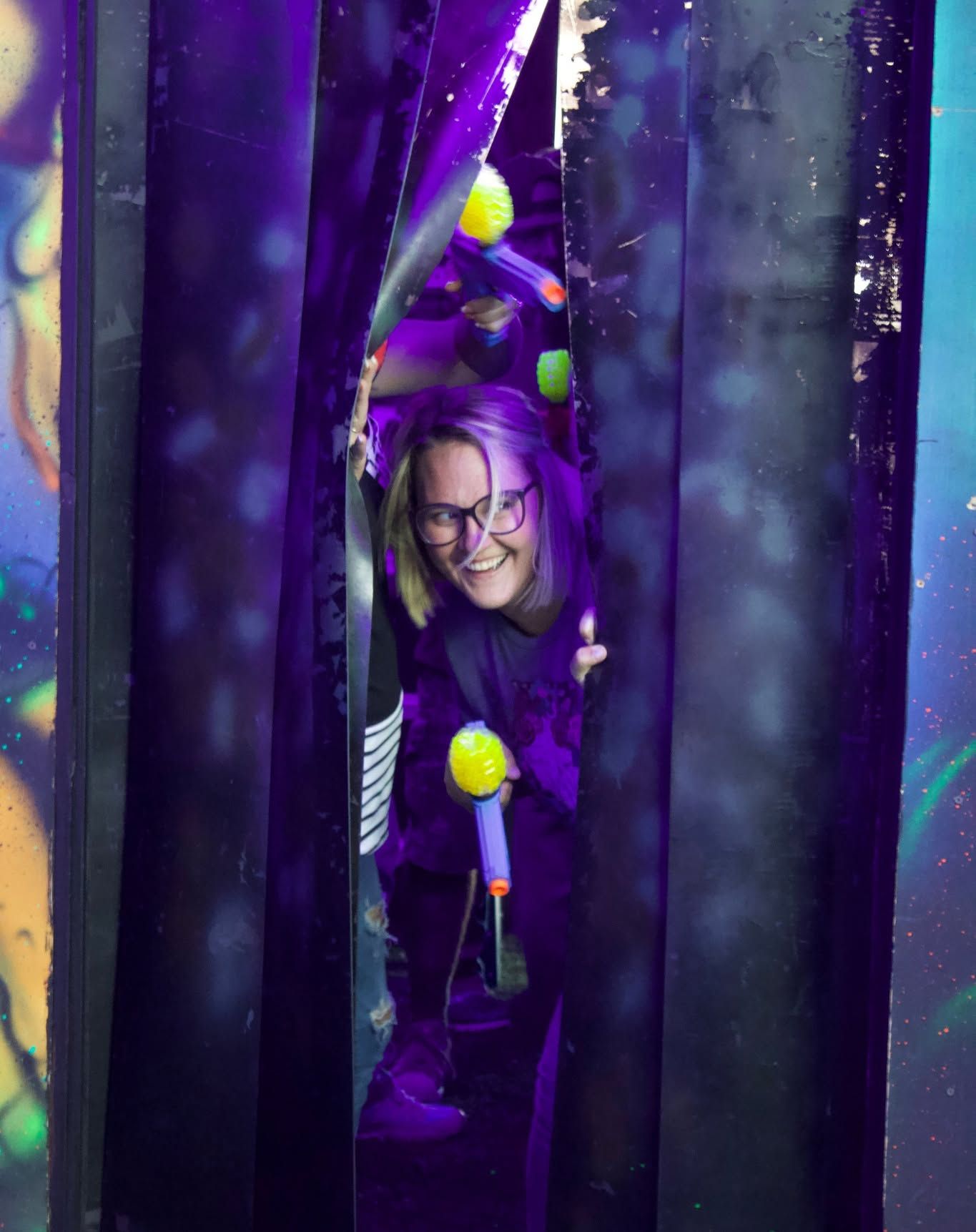A woman with purple hair and glasses is peeking out of a purple tunnel.