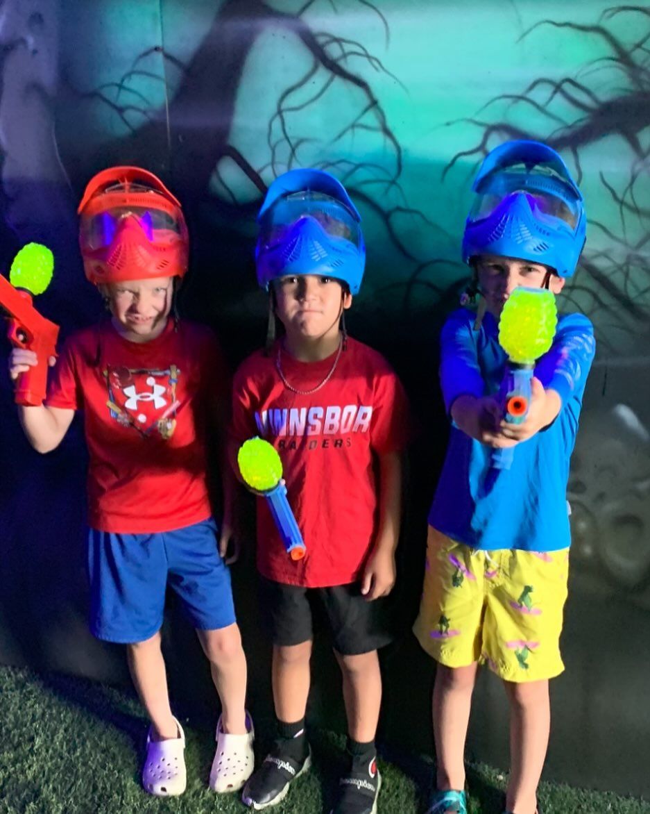 Three kids in glowing gear posing with toy blasters in a blacklight arena.