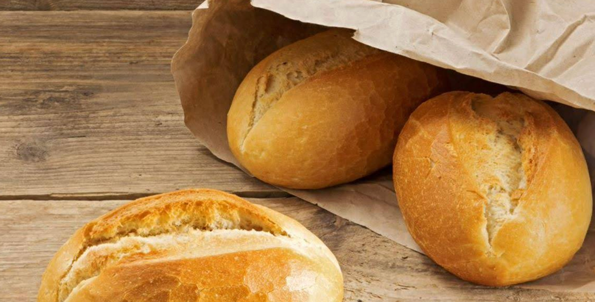Three golden-brown bread rolls, one in a brown paper bag, on a rustic wooden table.
