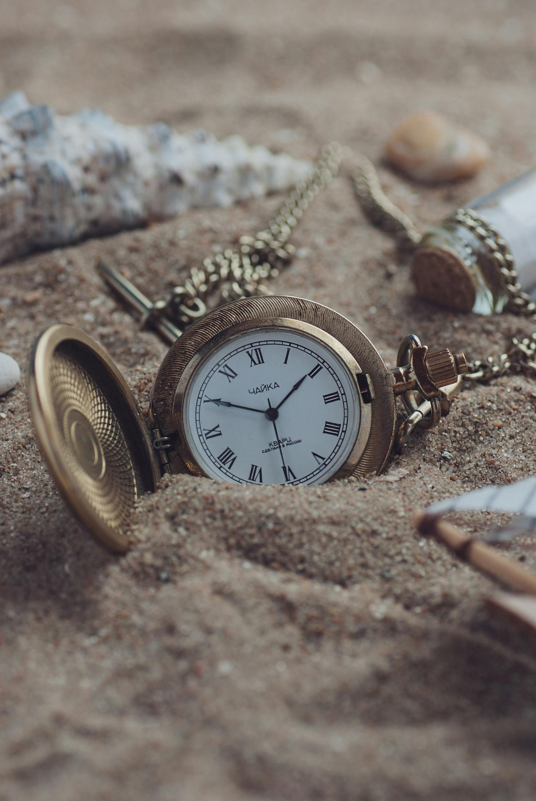 Gold pocket watch half-buried in sand with seashells; open lid.