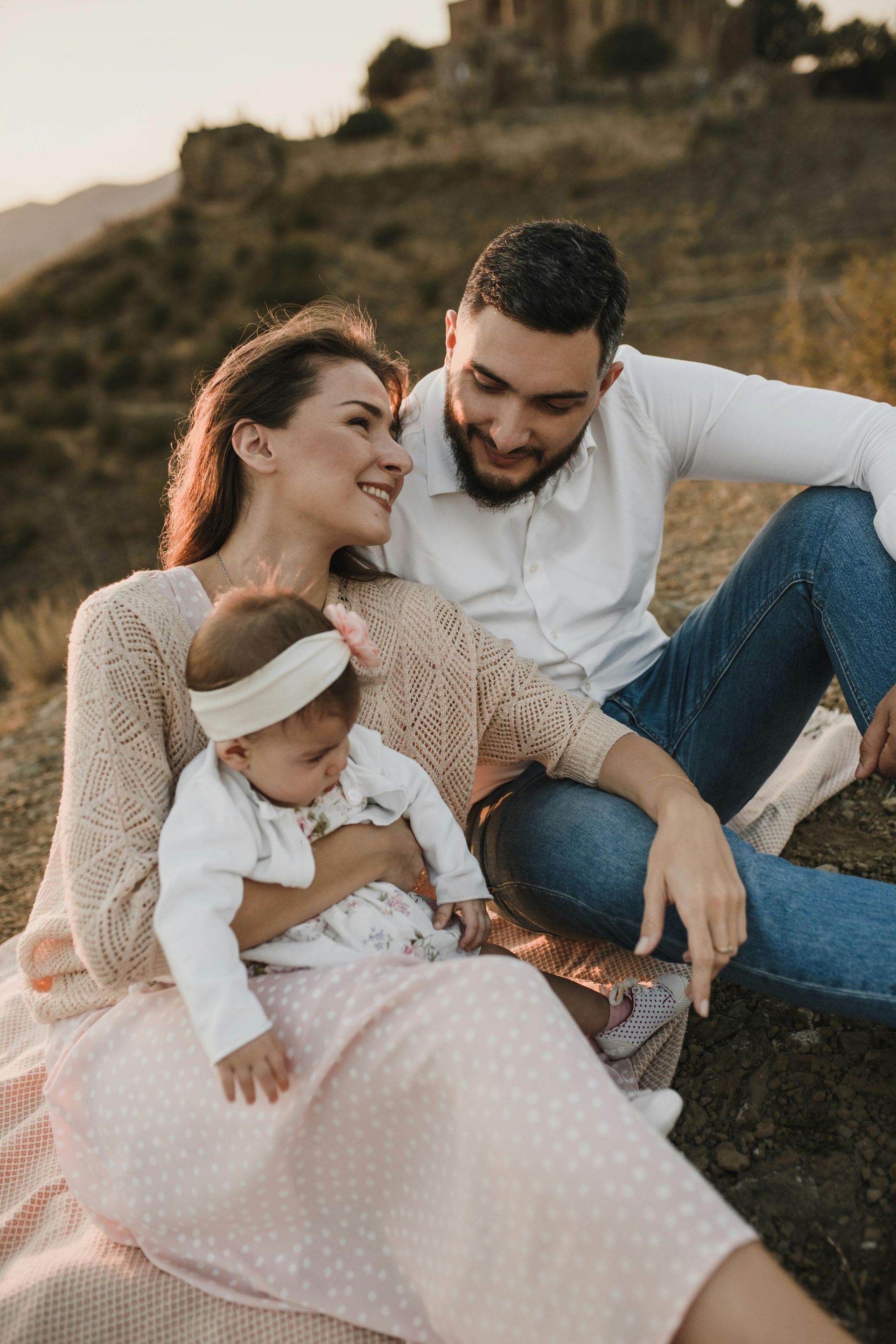 Family of three seated outdoors, mother holding baby, father smiling. Soft lighting.