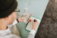 Person counting cash and writing on a notebook, sitting on a light blue bench.