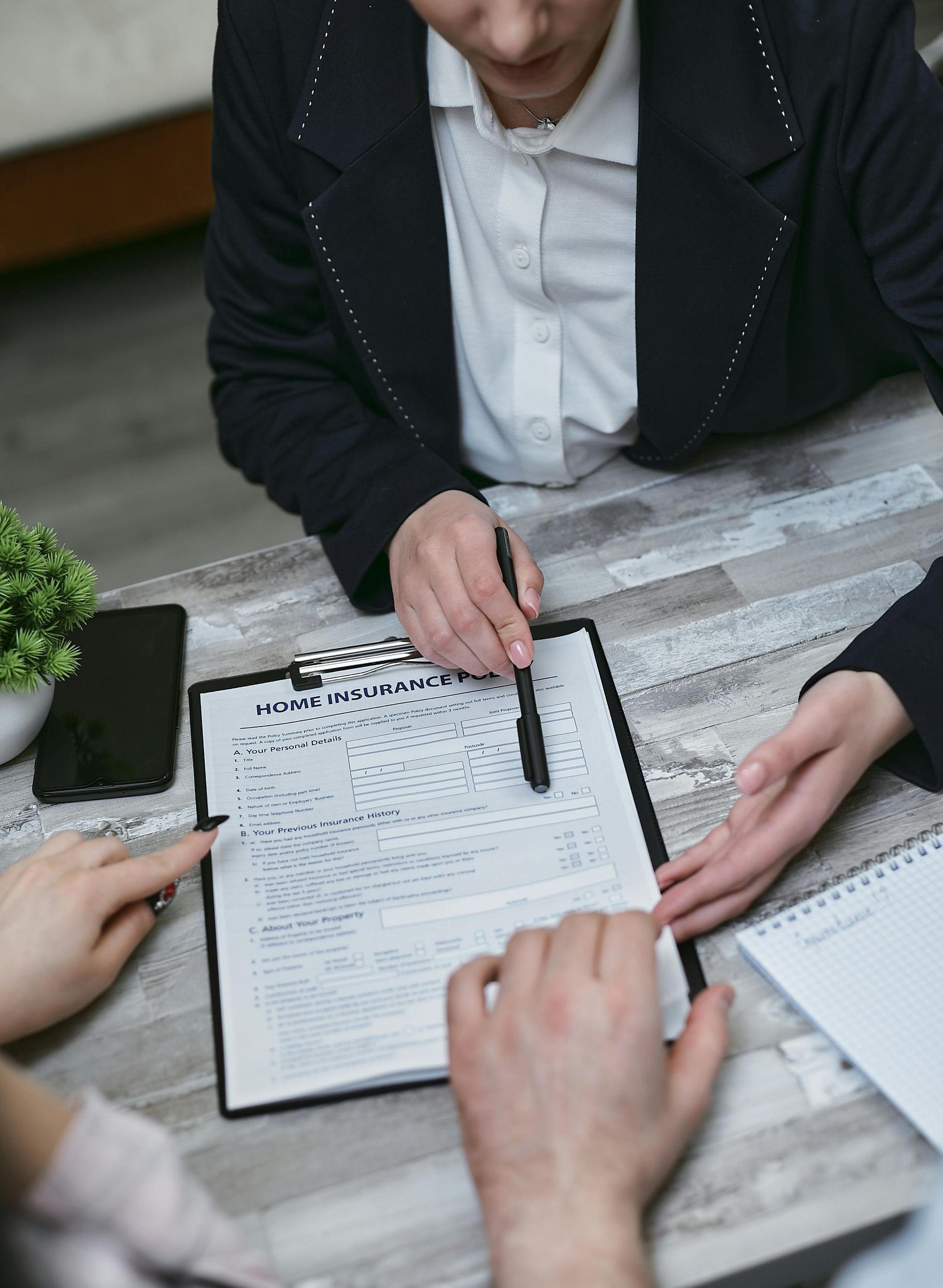 Person in black suit pointing at a contract on a clipboard. Two people are seated, observing the document.