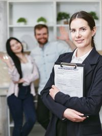 Real estate agent holding clipboard, smiling, with a blurred couple happily in background.
