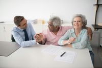 Man helping an elderly couple reviewing documents at a desk; man has arm around the man, all are smiling.