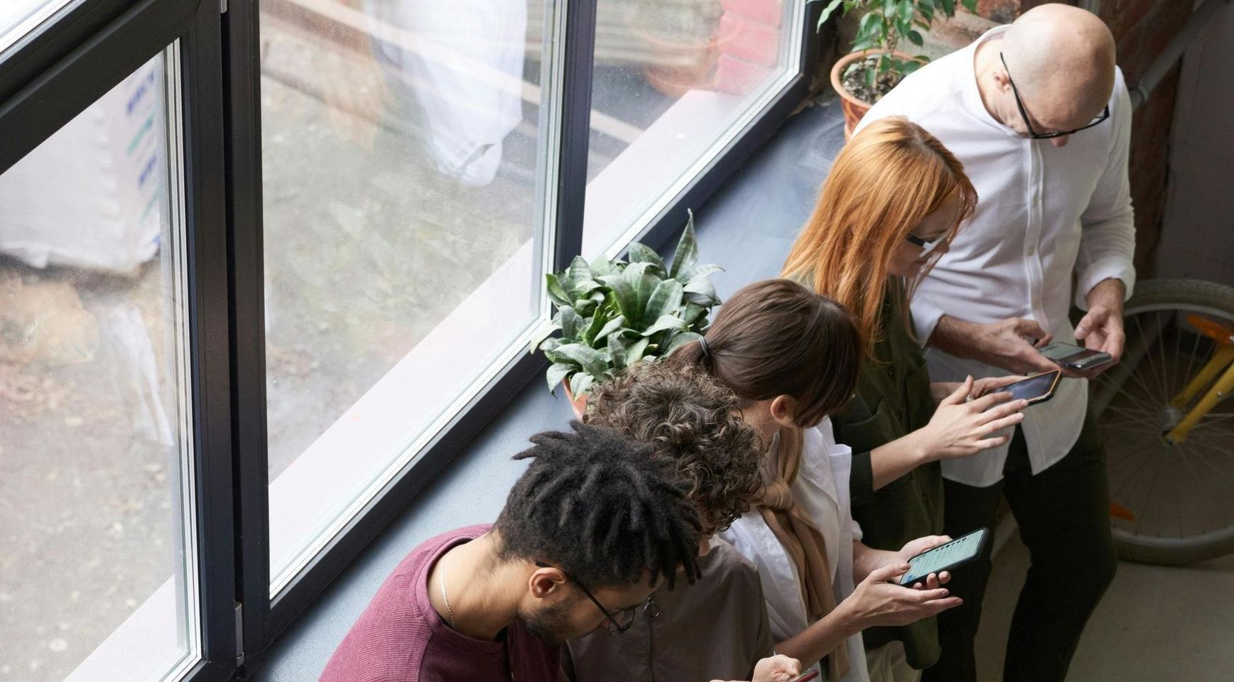 A group of people are standing in front of a window looking at their phones.