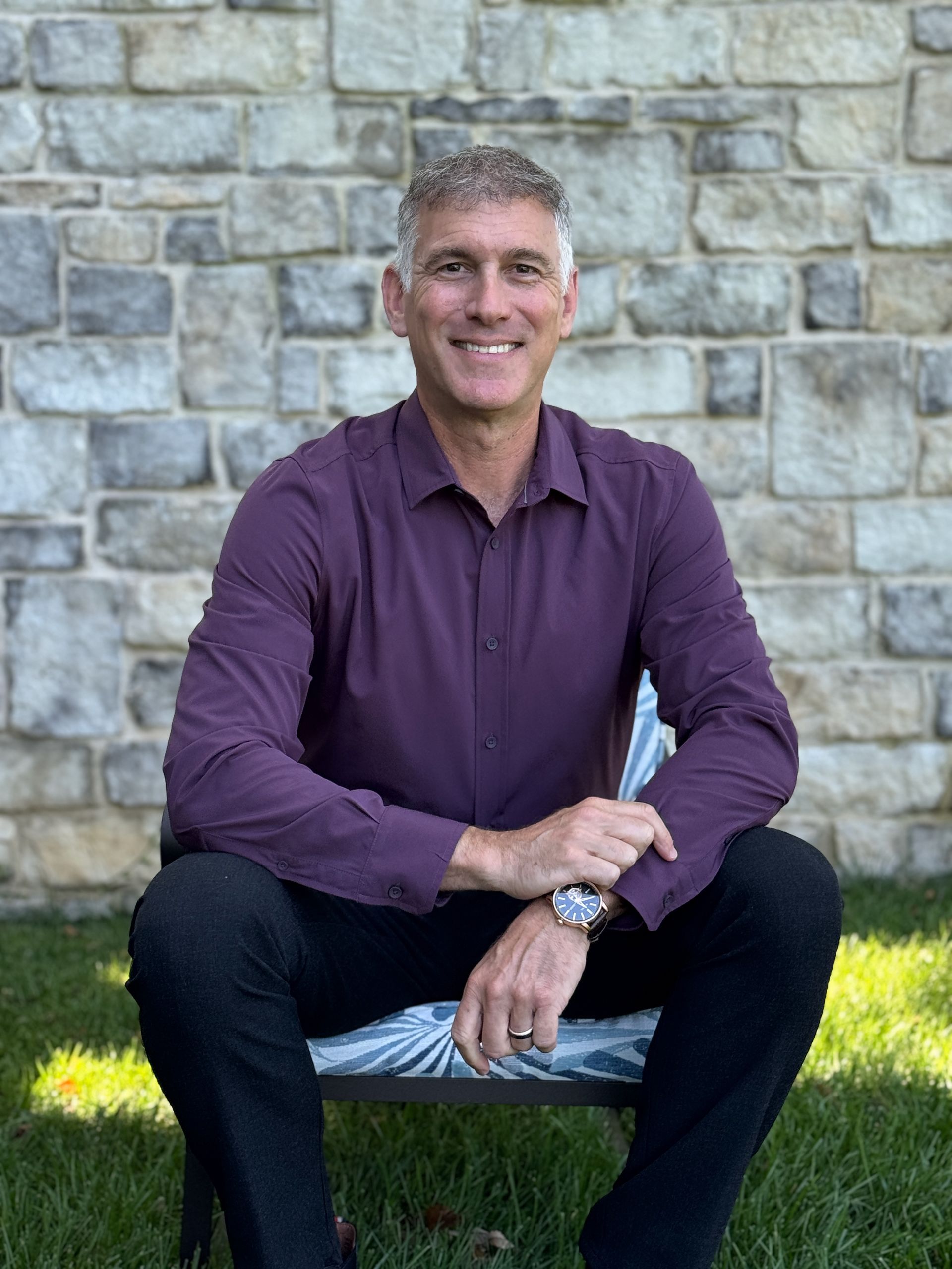 Man in a purple shirt seated outdoors against a stone wall, smiling at the camera.