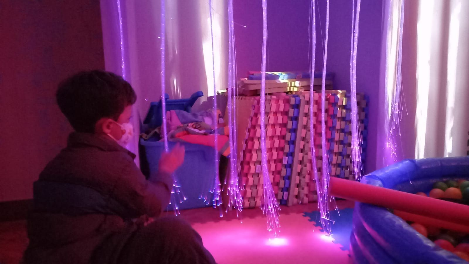 Un niño pequeño está sentado en una habitación con luces violetas y una piscina de pelotas.