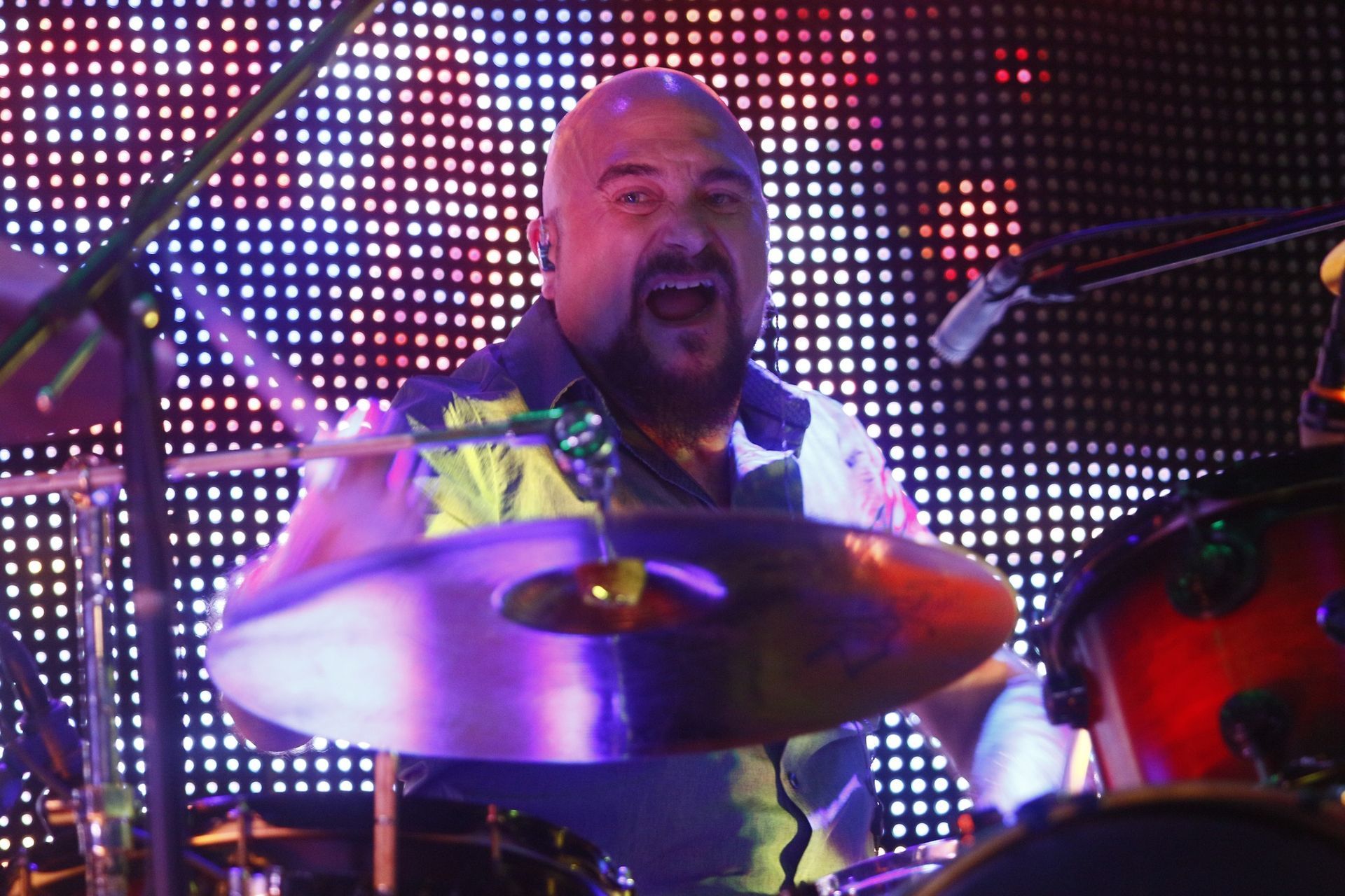 A bald man with a beard is playing drums in front of a colorful background.