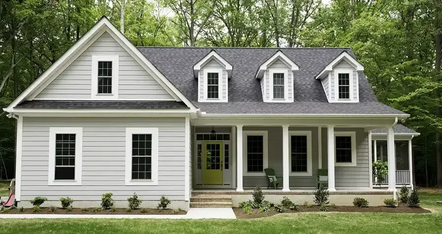 White two-story house with gray roof, dormer windows, and a covered front porch surrounded by trees