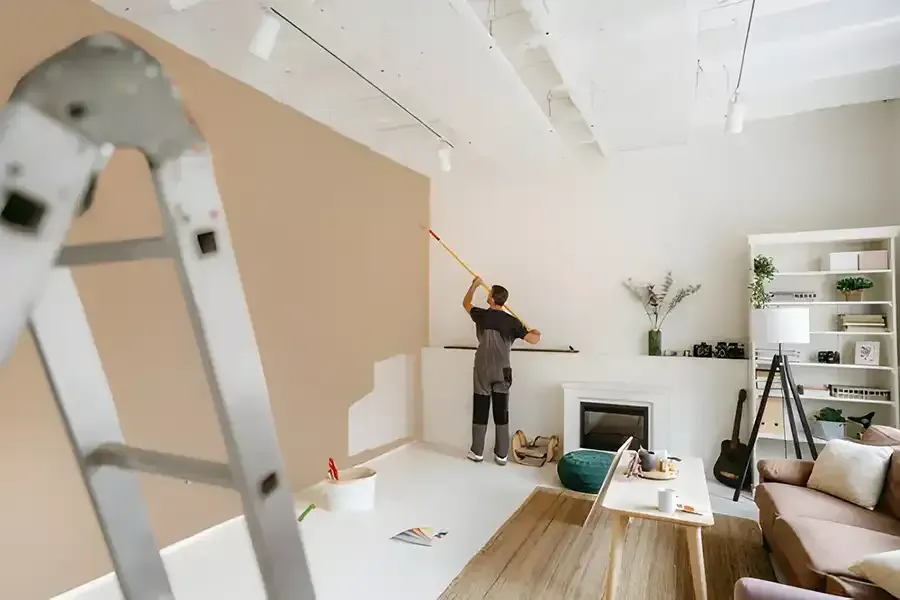 Person painting a beige wall in a bright living room with a ladder in the foreground