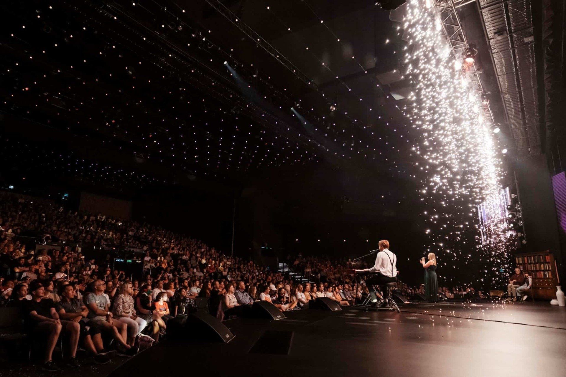 A man is standing on a stage in front of a crowd of people watching a fireworks display.