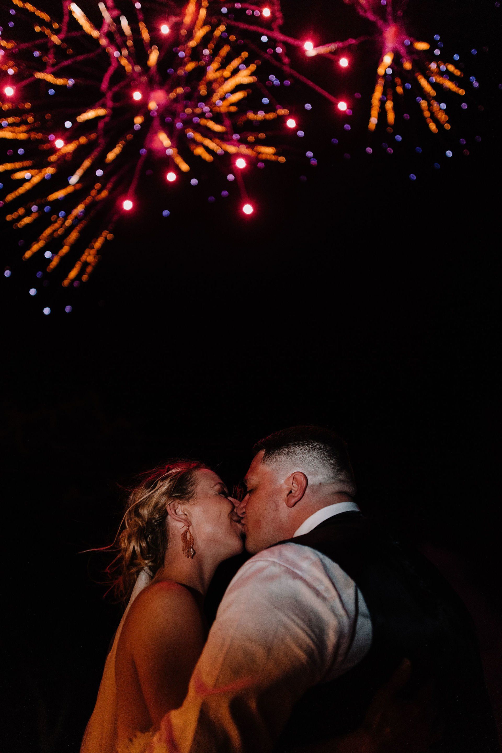 A bride and groom kissing in front of a fireworks display.