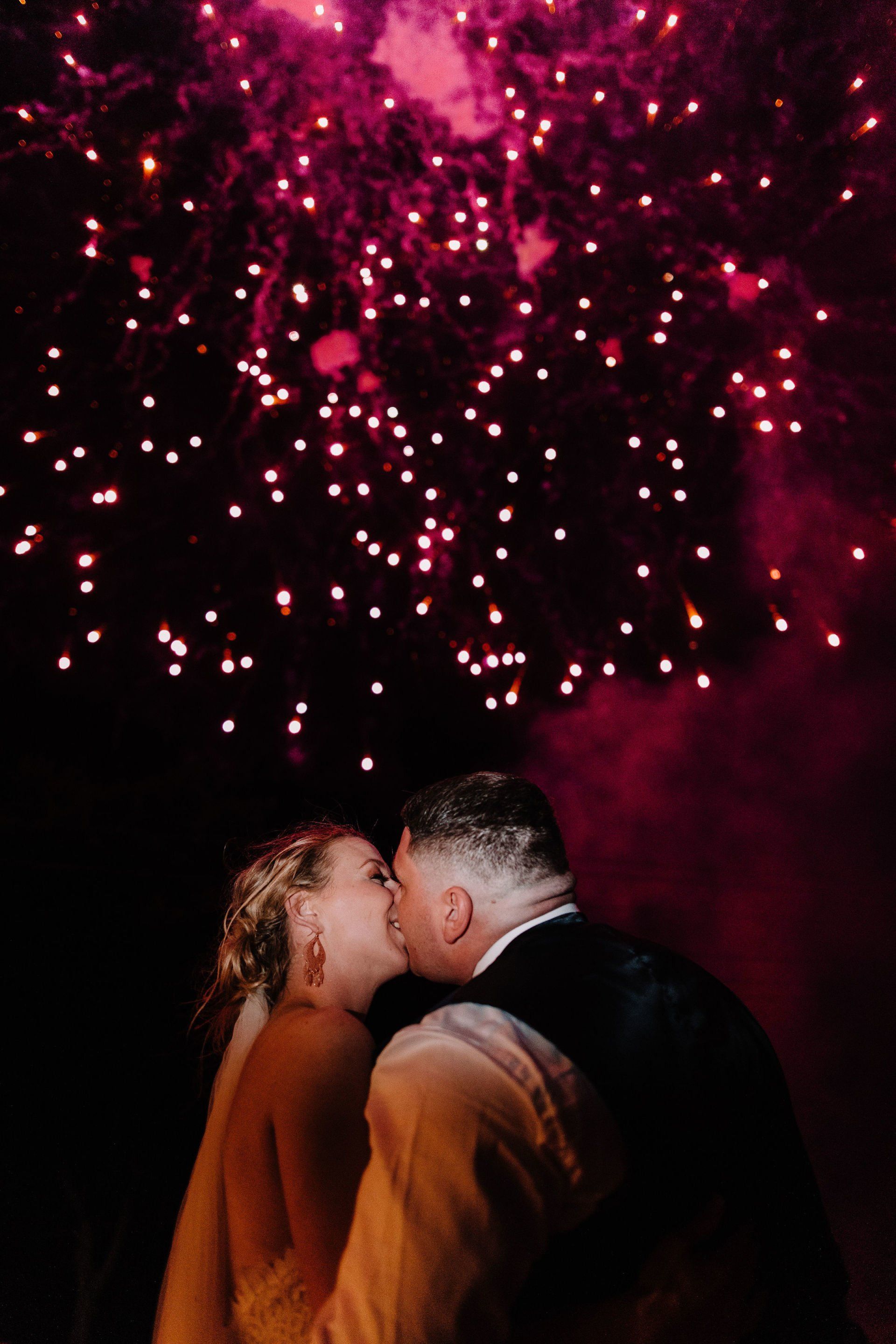 A bride and groom kissing in front of a fireworks display.