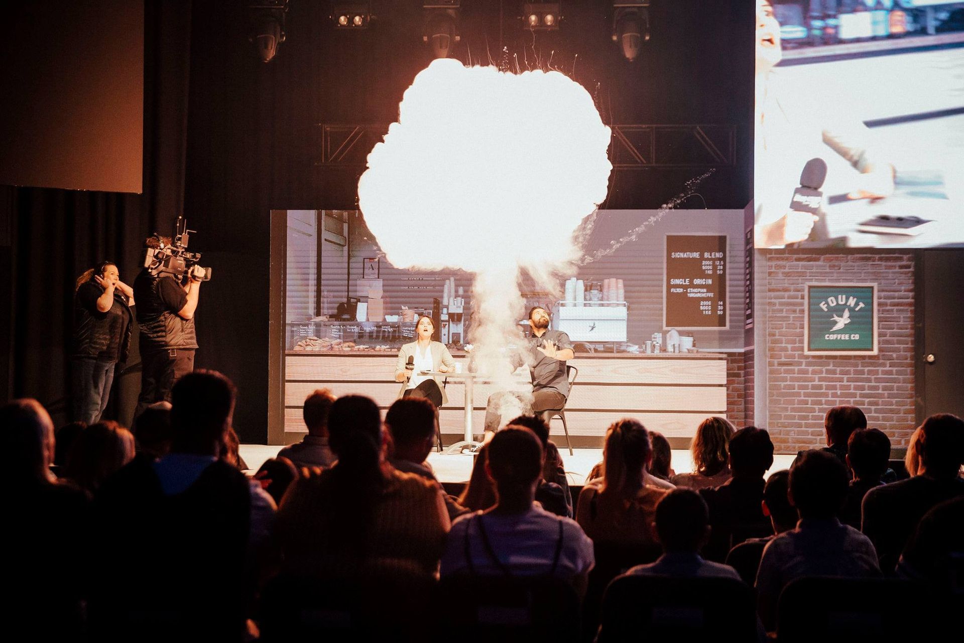 A group of people are sitting in front of a stage with fire coming out of it.