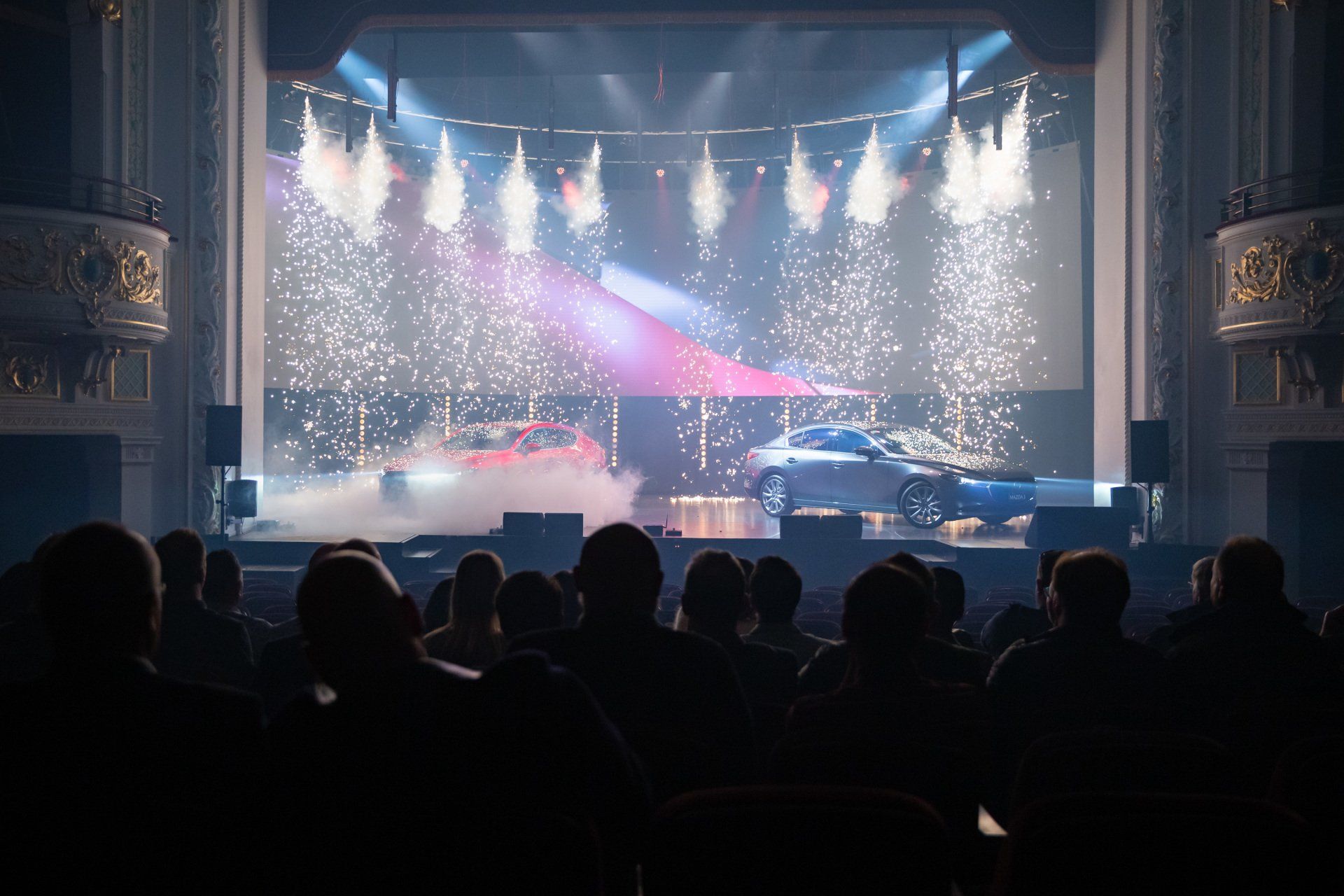 A group of people are sitting in front of a stage with a car on it.