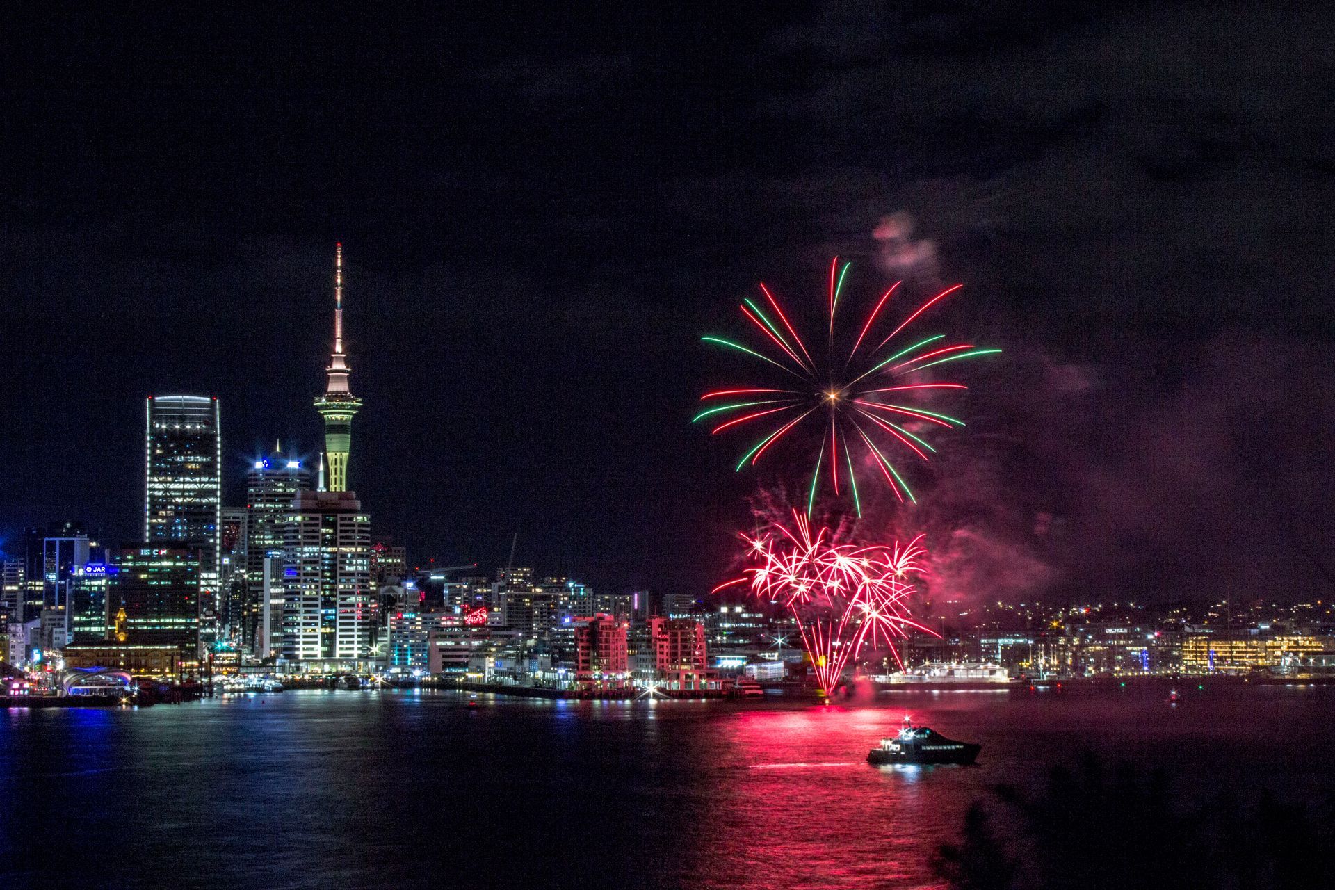 A fireworks display over a city with a boat in the foreground set over Auckland Harbour