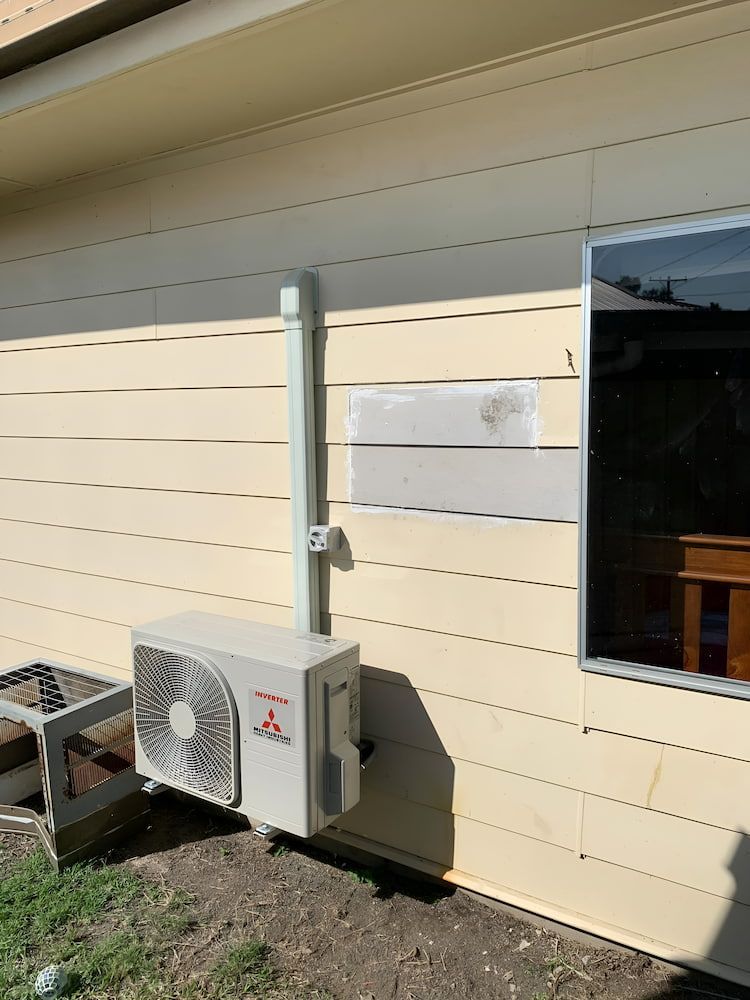 A White Air Conditioner Is Sitting On The Side Of A House Next To A Window — Levi's Electrics in Gladstone, QLD