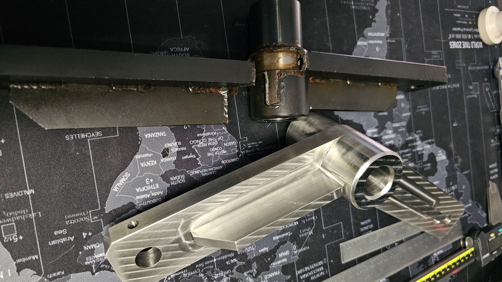 Machined metal parts with a black-and-white world map in the background. Tools are also present.