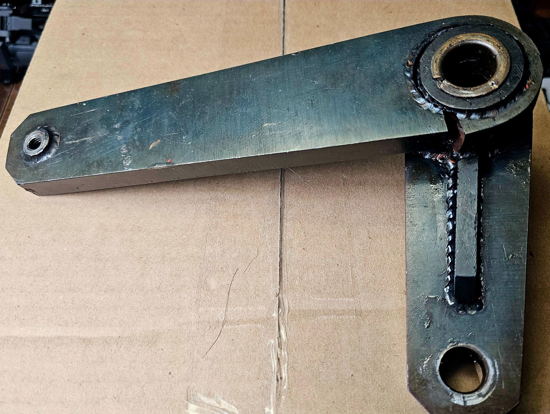 Metallic, L-shaped mechanical arm with a cylindrical bearing and bolt holes.