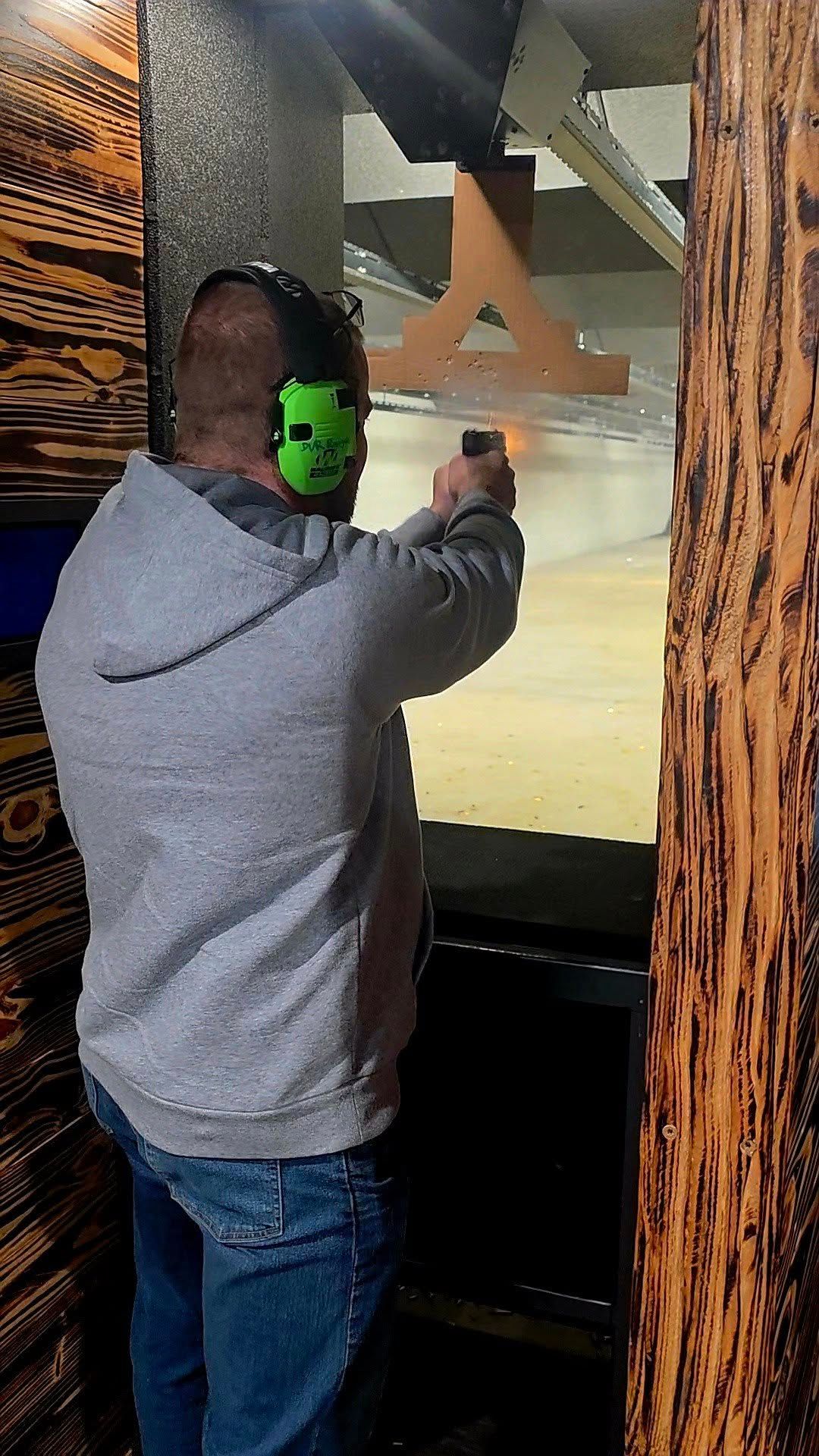 Man aiming a handgun at a shooting range, wearing ear protection and a gray hoodie.