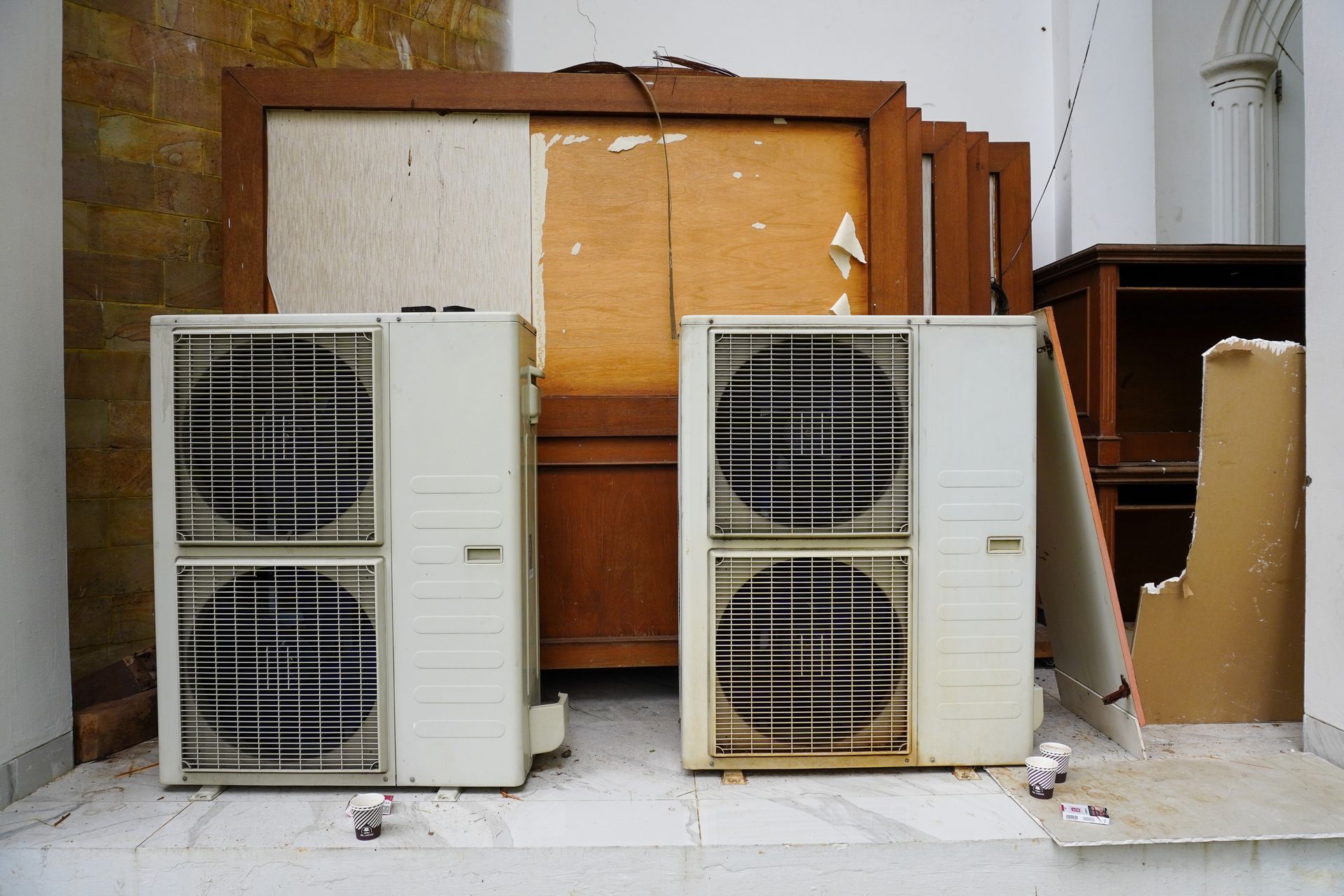 Two weathered air conditioning units on a dirty tiled floor with damaged wooden panels in the background.