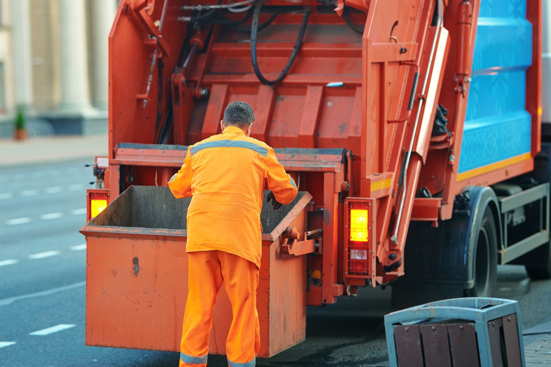 Garbage worker in orange uniform emptying trash bin into an orange garbage truck on a city street.