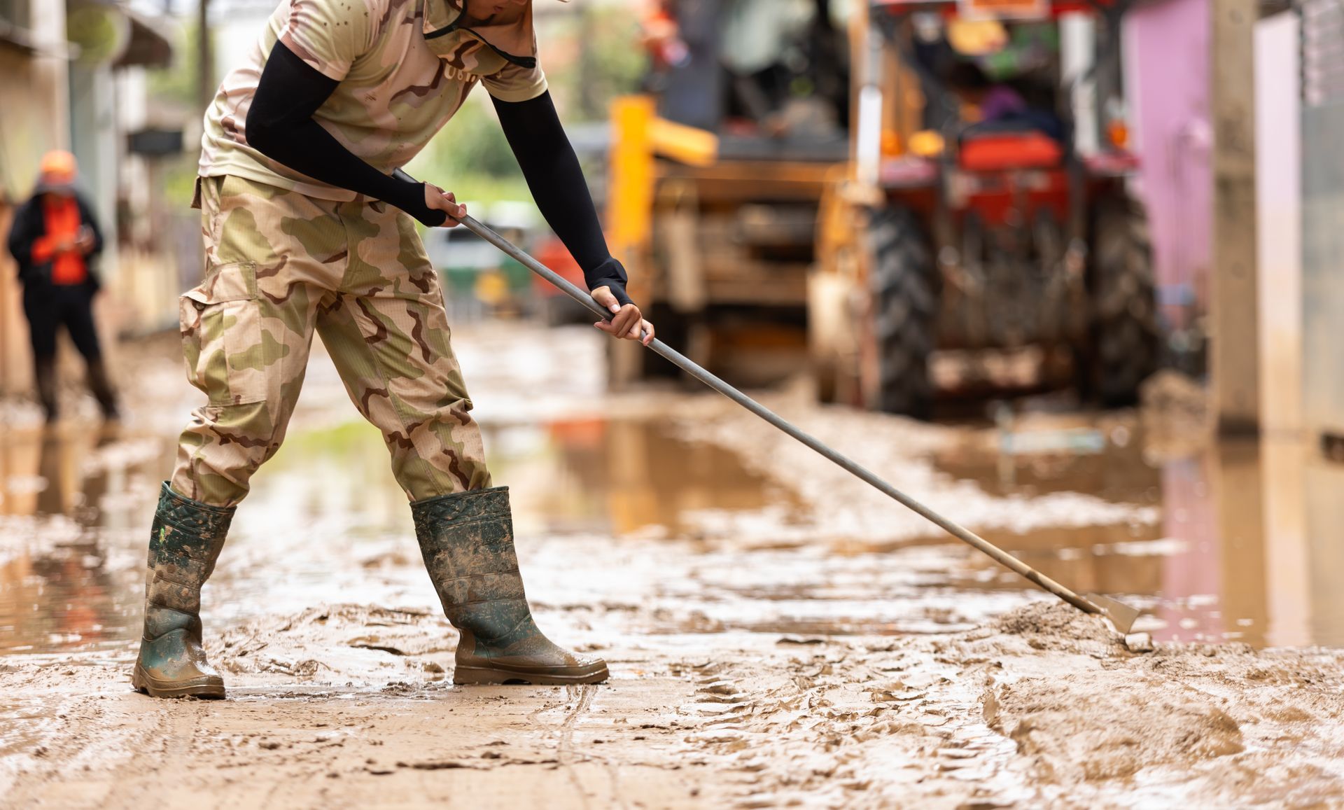 Person in camouflage clothes cleaning mud from a flooded street; tractor in the background.