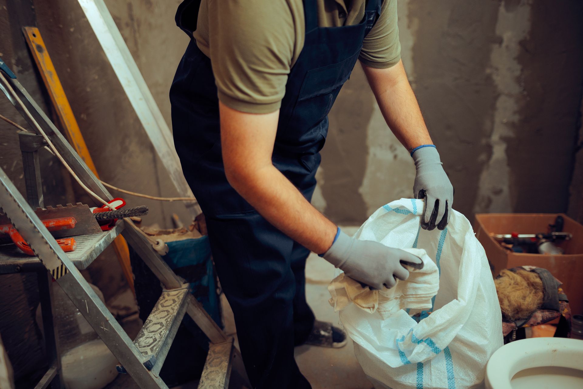 Person in overalls and gloves filling a bag with debris in a construction setting.