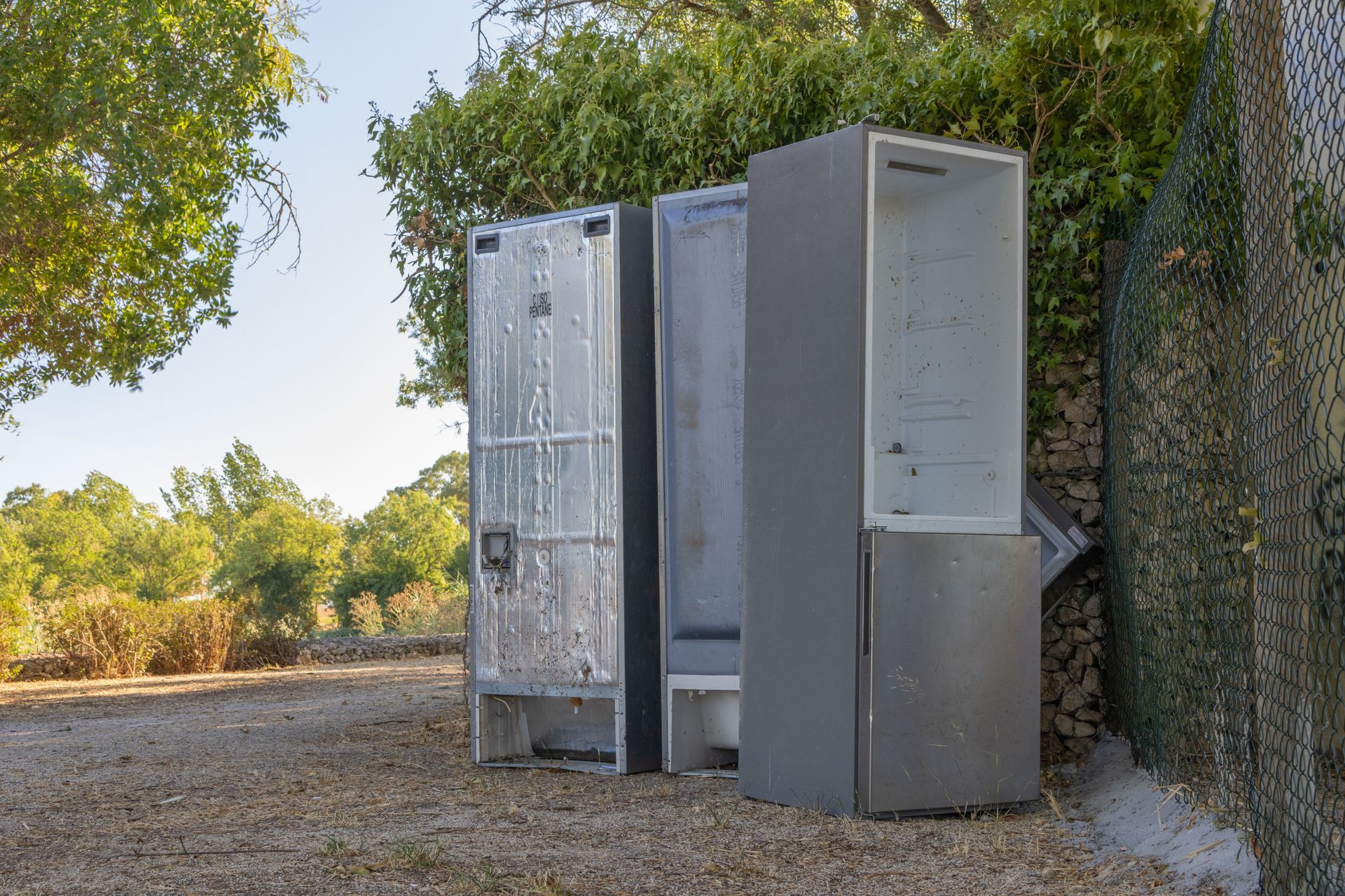 Three discarded metal storage lockers outdoors near a chain link fence and trees.