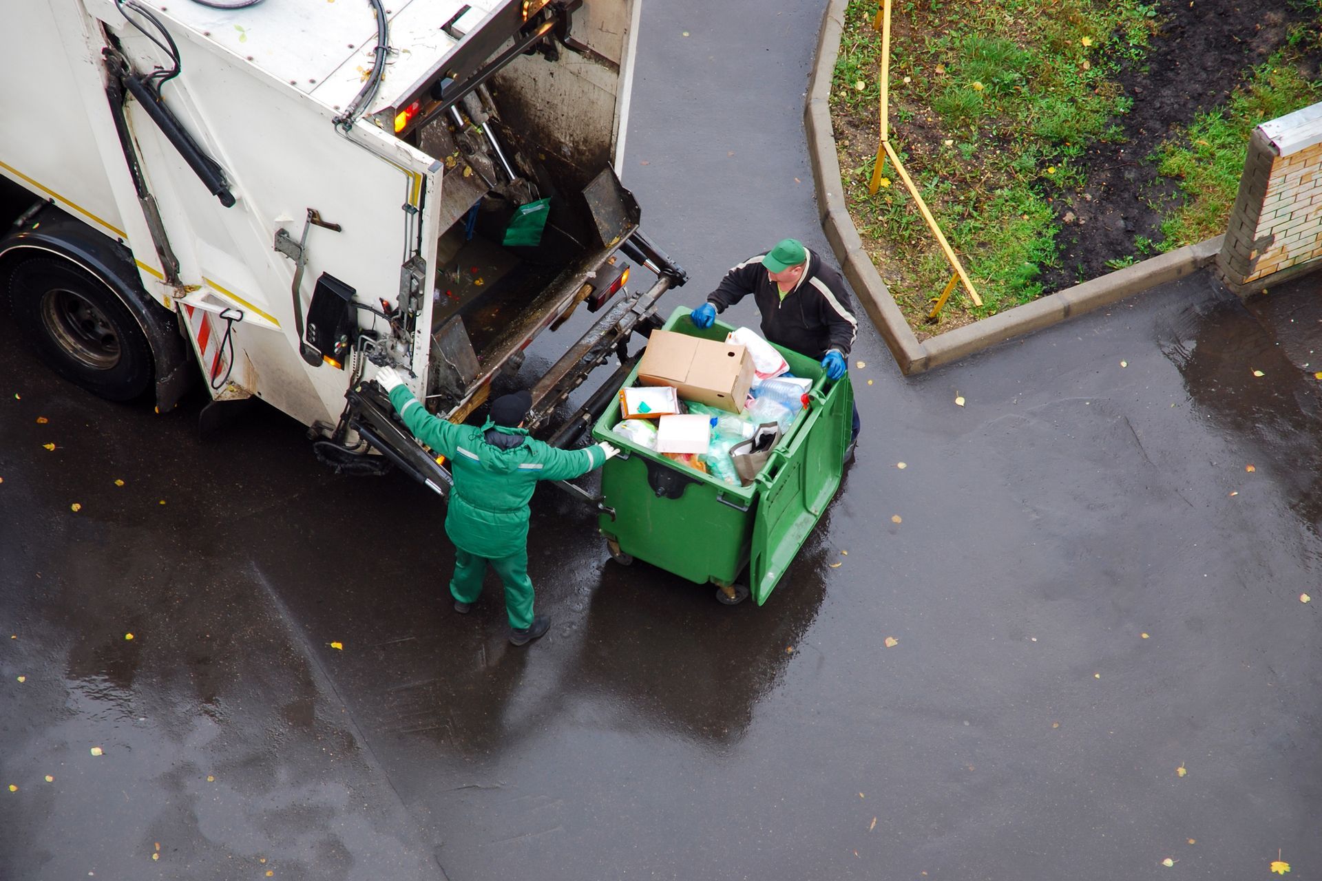Two sanitation workers emptying a full green dumpster into a garbage truck on a wet pavement.