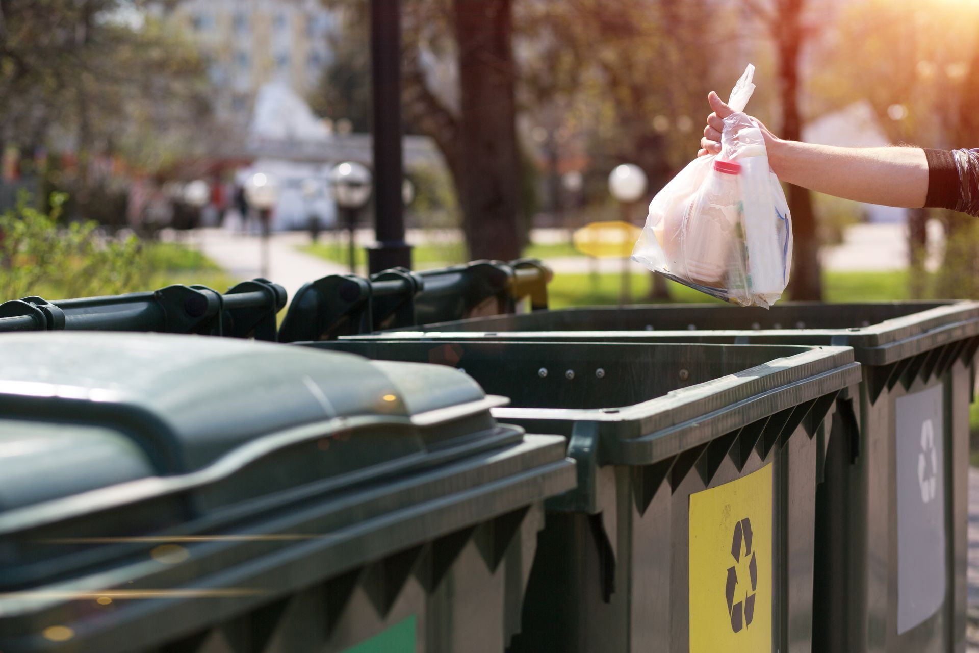 Person throwing trash bag into recycling bin outdoors.