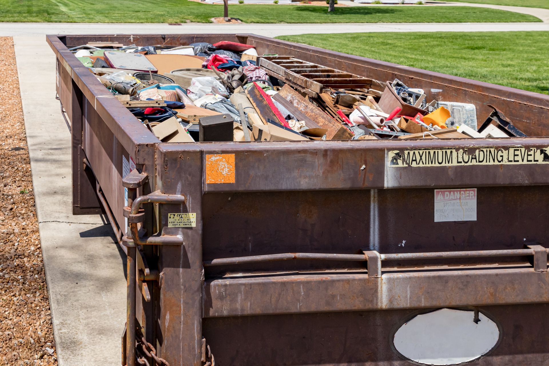 Dumpster overflowing with various discarded items, sits on concrete near a grassy area.