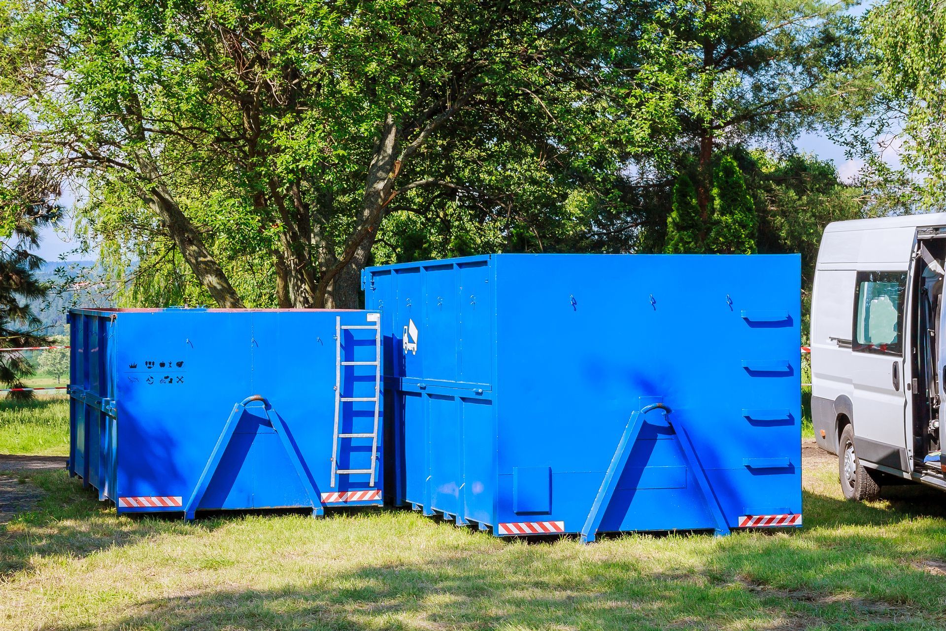 Two large blue industrial dumpsters outdoors with ladder and vehicle in background.