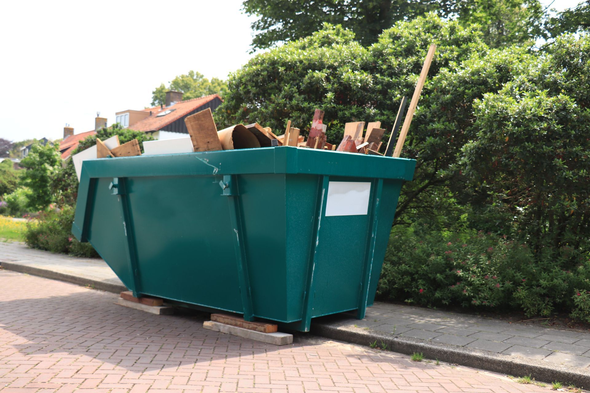 Green dumpster overflowing with debris on a brick sidewalk next to greenery.