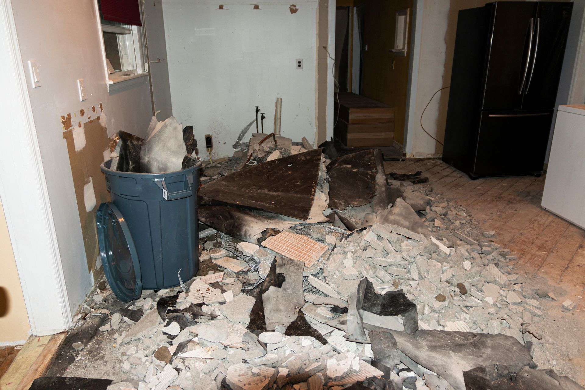 Demolition debris litters a room with a black refrigerator and blue trash can, near an open doorway.