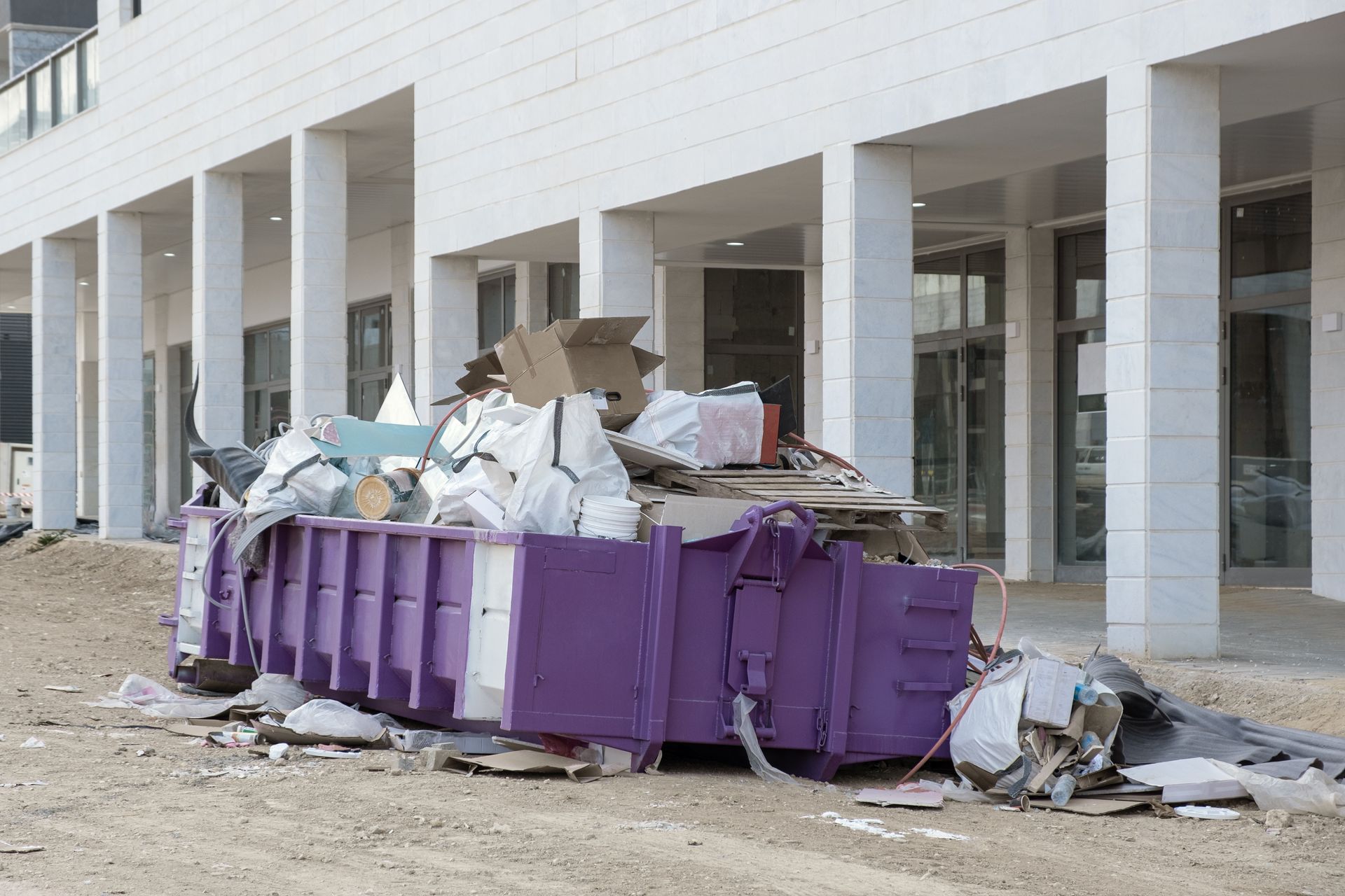 Purple dumpster overflowing with trash in front of a modern white building with columns.