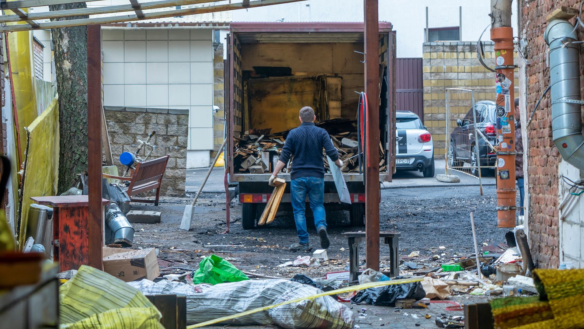 Man carrying wood into a truck in an alleyway; debris and vehicle visible.