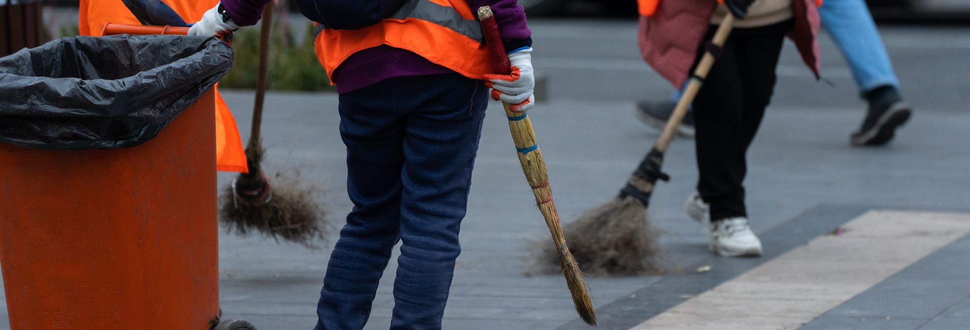 Street cleaners wearing safety vests sweep a road, orange trash can in view.