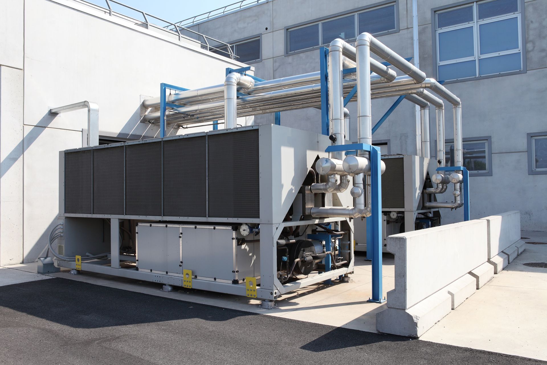 Industrial HVAC unit and piping outdoors beside a building, with a large gray equipment enclosure. Industrial HVAC unit and piping outdoors beside a building, with a large gray equipment enclosure.