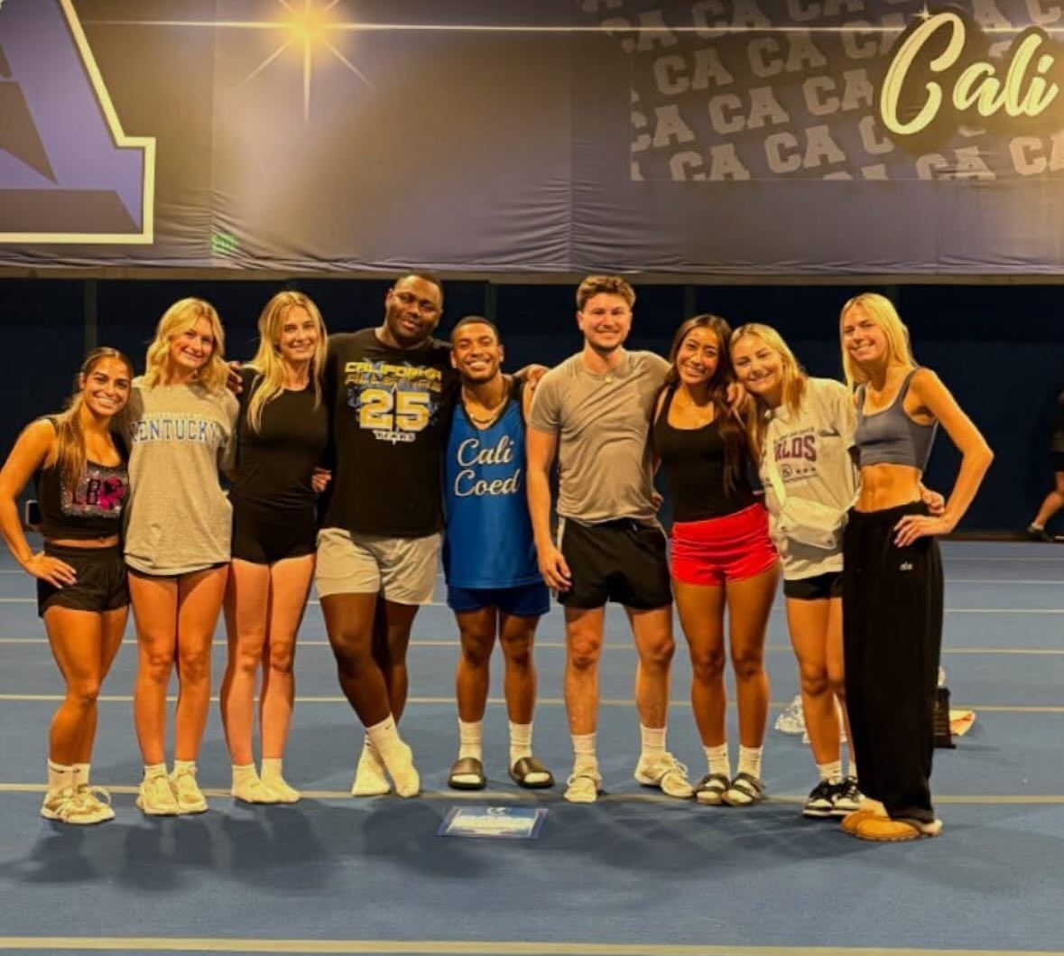 Group of cheerleaders posing in front of a banner. They are smiling, wearing athletic wear, and standing on a blue mat.
